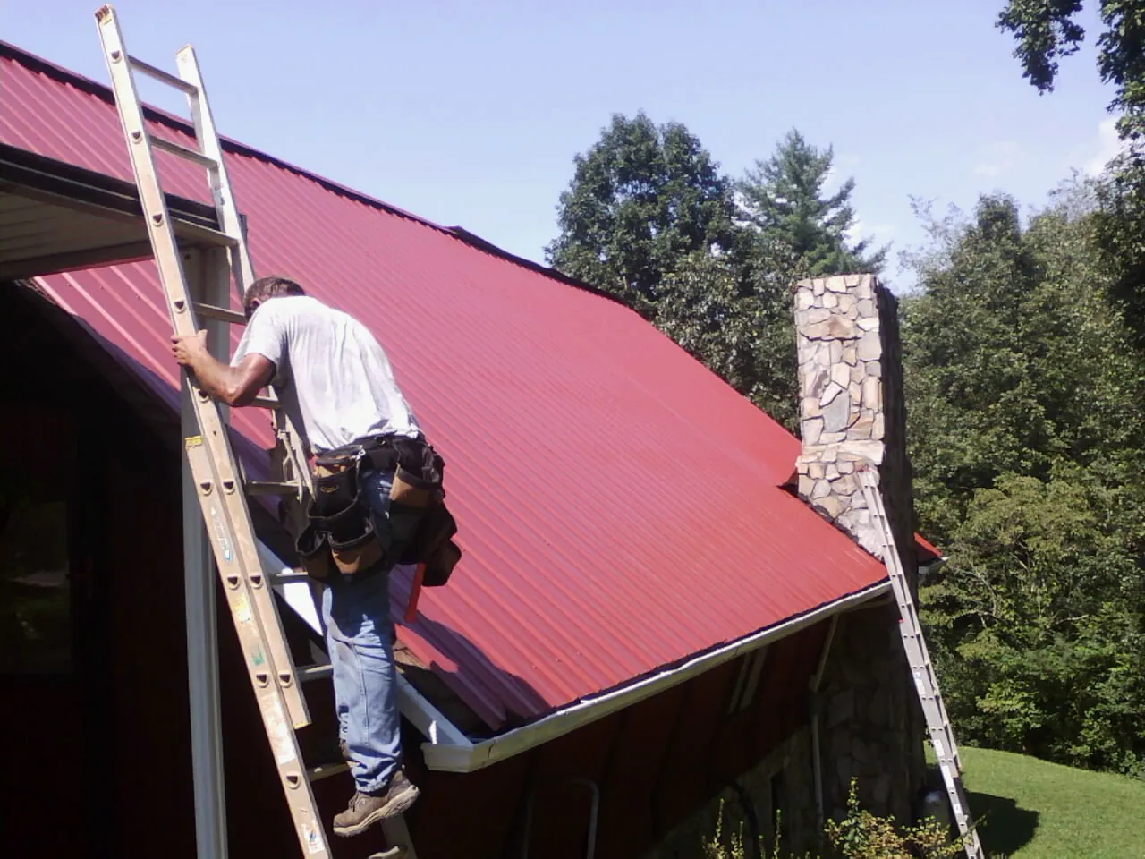 Roofer on a ladder beside a red metal roof and stone chimney — Soffit & Fascia Repair by Roofing Brothers in Dolphin